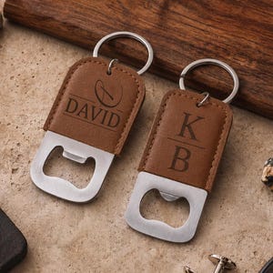 May include: Two brown leather bottle openers with silver metal rings. One opener is engraved with the name DAVID and a circular logo, while the other is engraved with the initials K and B separated by a horizontal line. Both feature a flat metal base.