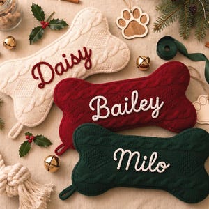 May include: Three bone-shaped pet stockings made of cable-knit fabric in cream, dark red, and dark green. Each stocking features a personalized name embroidered in a contrasting color: Daisy in red, Bailey in white, and Milo in white.
