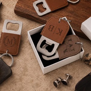May include: Three brown leather bottle opener keychains with metal hardware. One features a letter M in a laurel wreath, one has the initials KB, and one is engraved with the name Michael. They are presented in a white box with a black interior.