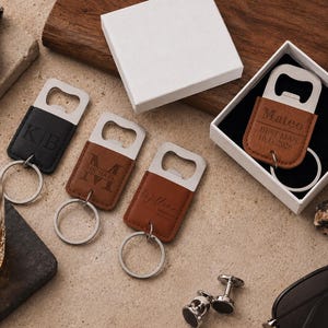 May include: Four rectangular bottle opener keychains with leather-like covers in black and brown. Three are laid out with engraved initials or names, while one is inside a white gift box with the text Mateo, BEST MAN, and 10.11.2026 engraved.