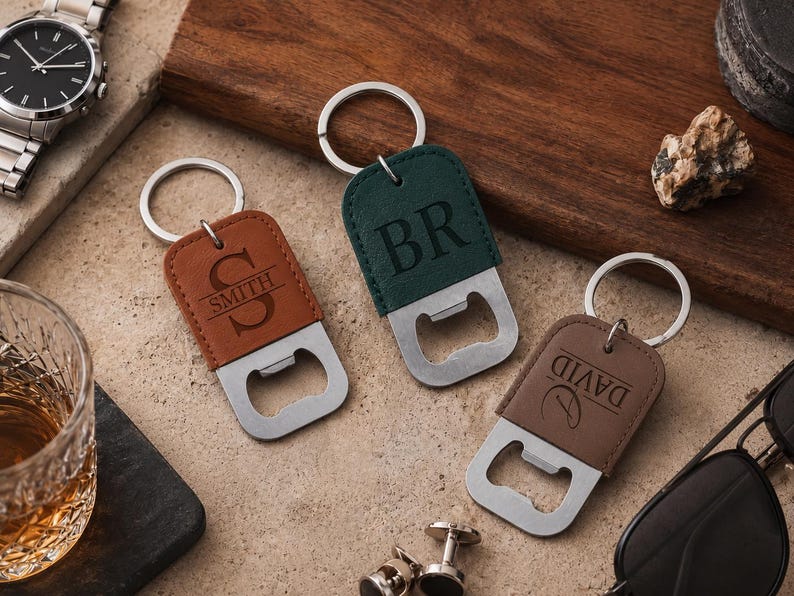 May include: Three rectangular bottle openers with leather-like covers in tan, dark green, and brown. Each features a silver metal ring and is engraved with personalized text: 'S SMITH', 'BR', and 'DAVID'. The openers have a flat metal base.