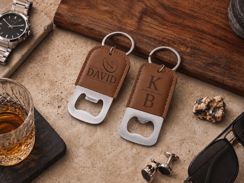 May include: Two brown leather bottle openers with silver metal rings. One opener is engraved with the name DAVID and a circular logo, while the other is engraved with the initials K and B separated by a horizontal line. Both feature a flat metal base.