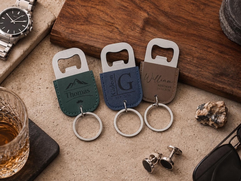 May include: Three metal bottle opener keychains with leather-like covers in dark green, blue, and brown. The green one features a mountain graphic and the name Thomas. The blue one displays the letter G and the name Gideon. The brown one shows William.