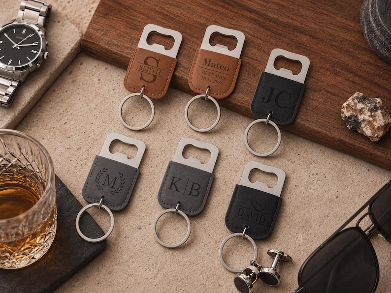 May include: Six rectangular metal bottle openers with leather-like covers in tan, black, and gray. Each features a silver split ring and custom laser-engraved text or monograms including Smith, Mateo, JC, M, KB, and David, arranged on a flat surface.