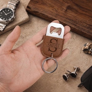 May include: A brown leather bottle opener keychain featuring a silver metal top and a circular ring. The leather portion is engraved with a large letter S and the text SMITH centered horizontally across the middle of the letter.