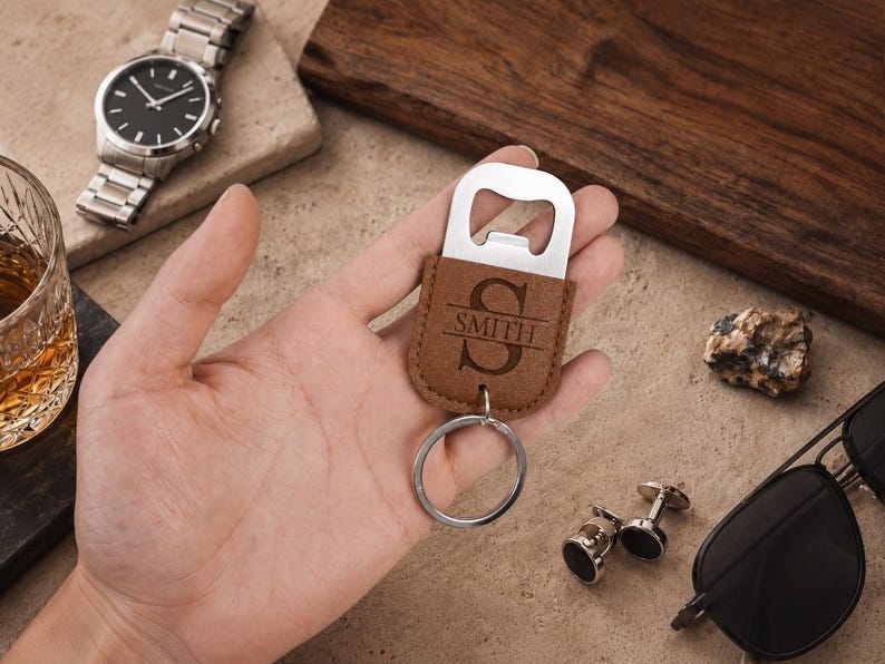 May include: A brown leather bottle opener keychain featuring a silver metal top and a circular ring. The leather portion is engraved with a large letter S and the text SMITH centered horizontally across the middle of the letter.