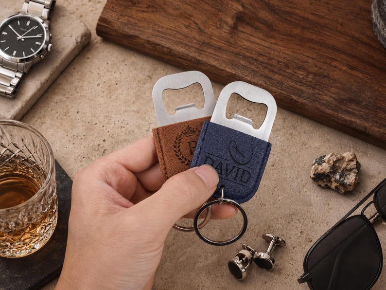 May include: Two metal bottle openers with leather-like covers in tan and navy blue. The tan cover features a crown and laurel wreath design, while the navy blue cover is embossed with the name DAVID and a coffee bean icon, each with a metal ring.