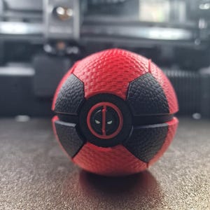 May include: A spherical object featuring a textured red and black pattern resembling a sports ball. The center displays a circular black emblem with a red border, showcasing the white-eyed mask icon of the character Deadpool in the middle.