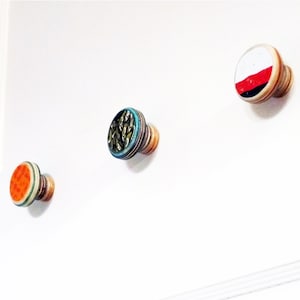 May include: Three decorative wall hooks made from wood with different patterns. The hooks are round and have a small wooden stem. One hook has a red, white, and black pattern, another has a green and blue pattern with gold accents, and the third has a multi-colored pattern with orange, white, and green.