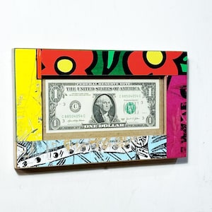 May include: A framed one-dollar bill. The frame is rectangular with a colorful design, including red, yellow, green, and blue sections. The bill features George Washington and the text "ONE DOLLAR".