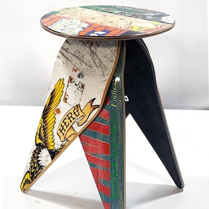 May include: A stool constructed from recycled skateboard decks featuring a circular seat with a multi-colored graphic print. The three legs are formed from curved skateboard sections displaying yellow eagle wing art, red text, and green Japanese characters.