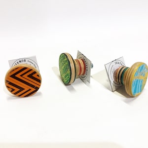 Peut inclure: Trois boutons de tiroir ronds en bois avec des motifs différents. Les boutons sont fabriqués à partir de plateaux de skate recyclés. Le premier bouton a un motif géométrique noir et orange. Le deuxième bouton a un motif vert, rouge et noir. Le troisième bouton a un motif bleu et jaune.