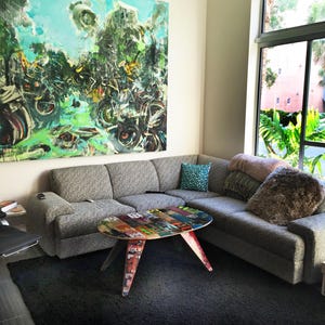 May include: A grey sectional sofa with cushions sits in a modern living room. A coffee table made from repurposed skateboards sits in front of the sofa. A large abstract painting hangs on the wall behind the sofa.