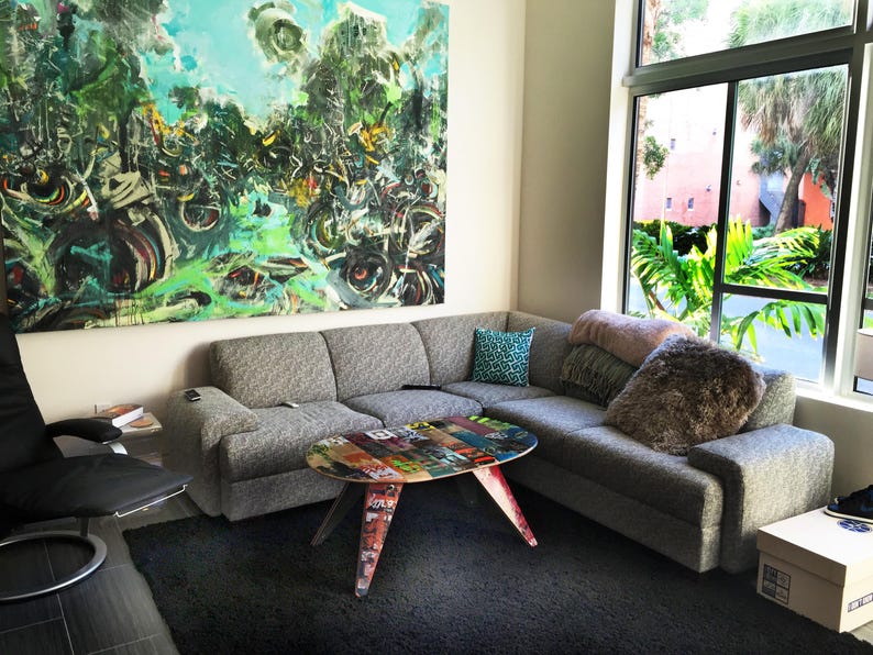 May include: A grey sectional sofa with cushions sits in a modern living room. A coffee table made from repurposed skateboards sits in front of the sofa. A large abstract painting hangs on the wall behind the sofa.
