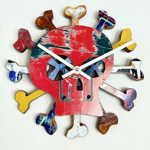 May include: A unique wall clock in the shape of a skull with crossbones, crafted from repurposed skateboard decks. The clock face is red, with white clock hands. The clock hands are reversible, white on one side and black on the other.