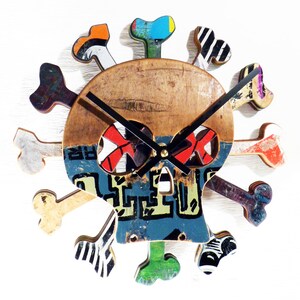 May include: A unique, handmade wall clock in the shape of a skull and crossbones. The clock face is made of wood and features a colorful, graffiti-style design with red, blue, green, and orange accents. The clock hands are black.