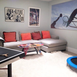 May include: A grey sectional sofa with red and orange throw pillows in a basement games room. A table tennis table with a bat is in the foreground. There is a basketball hoop poster on the wall, a poster with the text "High-Flying 360 Dunk" and a blue trampoline.