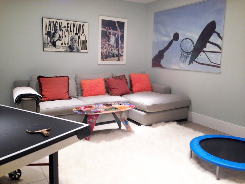 May include: A grey sectional sofa with red and orange throw pillows in a basement games room. A table tennis table with a bat is in the foreground. There is a basketball hoop poster on the wall, a poster with the text "High-Flying 360 Dunk" and a blue trampoline.