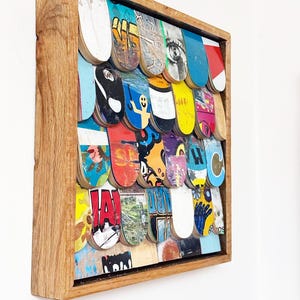 Può includere: Un'opera d'arte da parete incorniciata composta da tavole da skateboard colorate e riciclate. Le tavole sono disposte a griglia all'interno di una cornice in legno naturale. L'opera presenta una varietà di grafiche e colori, tra cui blu, rosso, giallo e bianco.