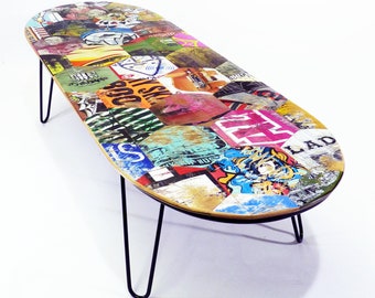 May include: A colorful oval coffee table made from a skateboard with black hairpin legs. The table top is covered in a collage of colorful stickers and graphics.