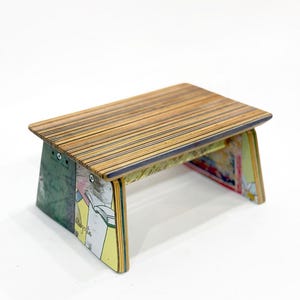 Puede incluir: Un pequeño taburete de madera hecho de tablas de skate recicladas. La parte superior del taburete está hecha de madera en capas con un acabado natural. Las patas del taburete están hechas de cubiertas de skate coloridas con diseños gráficos.