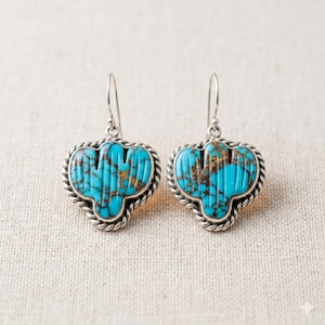 May include: A pair of drop earrings featuring bright turquoise stones with dark brown matrix patterns, carved into a cactus shape. The stones are set in silver frames with a twisted rope border and attached to silver shepherd hook ear wires.