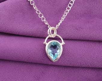 May include: A silver necklace with a pear-shaped, light blue gemstone pendant. The pendant is set in a silver frame and hangs from a delicate chain. The background is a solid purple fabric.