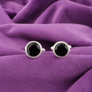 May include: A pair of silver cufflinks with large, round, black center stones. The cufflinks are set against a purple velvet background, creating a contrast between the shiny metal and the dark stones.