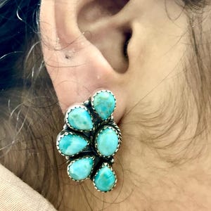 May include: A cluster earring featuring turquoise teardrop-shaped stones set in a silver-toned metal. The stones are a vibrant blue-green color, and the metal has a detailed, rope-like border. The earring is shown on an ear.