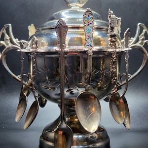 May include: A silver-colored serving bowl with a lid, adorned with a bird-shaped handle. The bowl is surrounded by several spoons hanging from the sides. The spoons have ornate handles and engraved details. The bowl sits on a pedestal base.