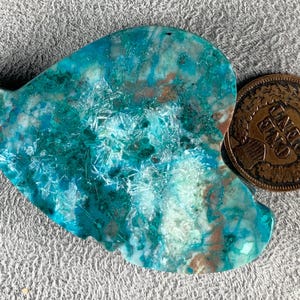May include: A heart-shaped, polished stone with a vibrant turquoise and teal color palette, accented with brown and white inclusions. A copper-colored one-cent coin rests beside the stone, providing a size reference. The stone's surface has a crystalline texture.