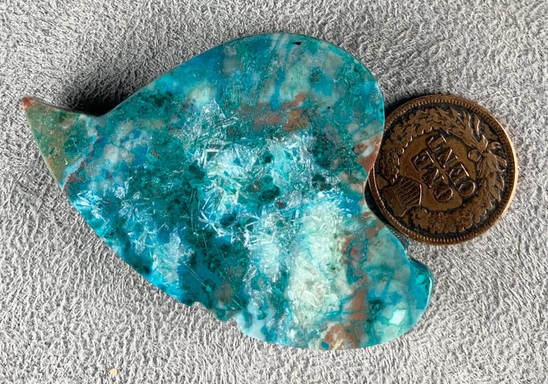 May include: A heart-shaped, polished stone with a vibrant turquoise and teal color palette, accented with brown and white inclusions. A copper-colored one-cent coin rests beside the stone, providing a size reference. The stone's surface has a crystalline texture.