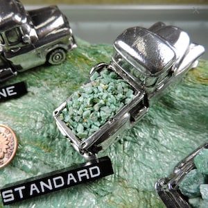 May include: Miniature silver truck models with open beds filled with small, light green stones. The trucks sit on a textured, green surface. The word "STANDARD" is visible on a black label.
