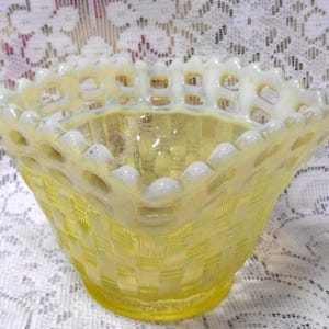 May include: A yellow glass bowl featuring a woven basket-weave pattern on the exterior and a white, scalloped, lattice-style rim. The glass has a translucent quality with a smooth, rounded base and a wide, open top with a decorative edge.