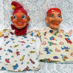 May include: Two vintage hand puppets with painted faces and fabric clothing. One puppet wears a red hat and bow tie, the other a red cap. Their cream-colored shirts are patterned with colorful cartoon figures.