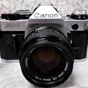 May include: A silver and black Canon AE-1 Program 35mm film camera with a black lens and strap. The Canon logo is visible on the top of the camera. The lens reads "Canon Lens FD 50mm 1:1.4".