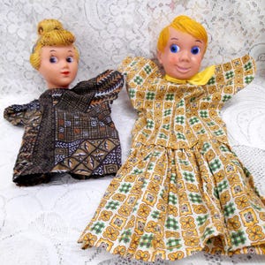 May include: Two vintage hand puppets. One puppet has blonde hair and wears a brown patterned dress. The other puppet has blonde hair and wears a yellow, green, and white patterned dress. The puppets are on a white lace surface.