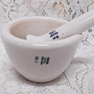 May include: A white ceramic mortar and pestle set. The mortar is bowl-shaped with a wide opening, and the pestle is cylindrical. The set has markings that read "GOODS U.S.A." and "23-1".