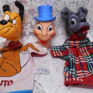 May include: Three vintage Disney character hand puppets: Pluto, Jiminy Cricket, and a gray dog. Pluto is yellow with a red collar and a shirt that says "PLUTO". Jiminy Cricket wears a blue top hat. The gray dog wears a plaid shirt.