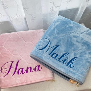 Puede incluir: Dos alfombras de oración personalizadas de felpa. Una es rosa claro con el nombre "Hana" bordado en morado. La otra es azul claro con "Malik" bordado en azul. Ambas tienen un patrón floral.