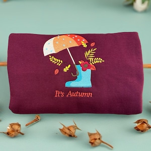 May include: A burgundy sweatshirt with an embroidered autumn design. The design includes a colorful umbrella, a blue boot with a flower, and falling leaves. The text "It's Autumn" is embroidered below the design.