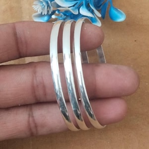 May include: A set of three silver bangles with a polished finish. The bangles are round and smooth, reflecting light. A blue and white floral accent is visible in the background. The bangles are displayed on a brown surface.