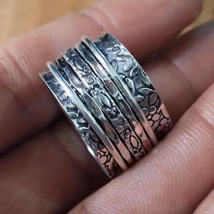 May include: A silver spinner ring with multiple bands. The bands feature intricate floral and leaf patterns, creating a textured and detailed design. The ring is shown close-up, highlighting the craftsmanship and the contrast between the polished and etched surfaces.