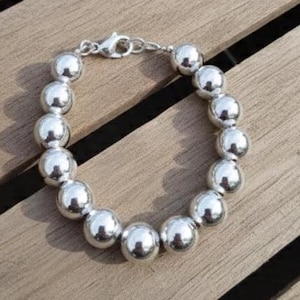 May include: A silver beaded bracelet with large, round beads. The bracelet is made of shiny, polished metal and is connected by a small clasp. The bracelet is displayed on a wooden surface.