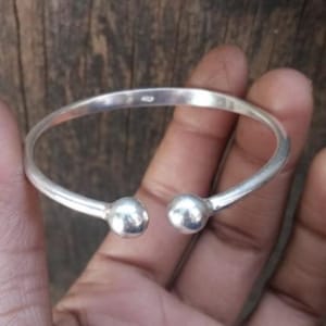 Könnte beinhalten: Ein silberfarbenes Armreif-Armband mit zwei kugelförmigen Endkappen. Das Armband wird in einer Hand vor einem Holzgrund gehalten. Das Armband hat ein schlichtes, elegantes Design.