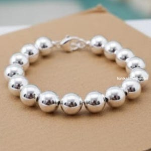 May include: A silver bracelet featuring large, round beads strung together. The beads have a polished, reflective surface. The bracelet is secured with a silver clasp. The jewelry is displayed on a brown surface.