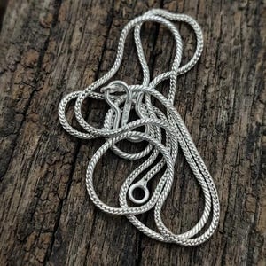 May include: A silver-coloured snake chain necklace featuring a textured, woven metal link design. The necklace includes a hook-style clasp mechanism and a circular loop closure, showcasing a polished metallic finish throughout the entire chain.