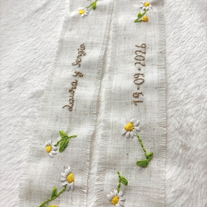 Op de afbeelding: Twee off-white linnen linten met geborduurde madeliefjes en groene bladeren. Eén lint heeft de tekst "Laura y Sergio", de andere leest "19-09-2026" in bruine draad. De linten zijn geplaatst op een witte, getextureerde ondergrond.