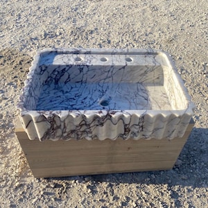 Puede incluir: Un lavabo rectangular de mármol con base blanca y vetas púrpuras prominentes. El borde frontal del lavabo presenta un diseño ondulado y festoneado. La parte superior incluye tres orificios pretaladrados para grifería y un desagüe central.