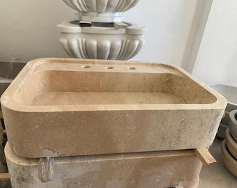 May include: A rectangular, light brown stone sink with three tap holes. The sink is stacked on top of another similar stone structure. A decorative marble element is visible in the background.